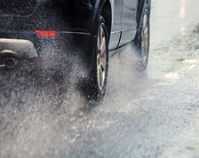 A dark SUV drives away from the camera through puddles of water, splashing the water into the air.